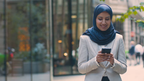 Muslim-Businesswoman-Checking-Messages-On-Mobile-Phone-Standing-Outside-Office-In-City-3